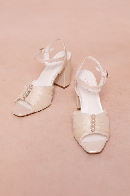 The Piper Pearl Heel in Ivory by Ivy City Co features elegant ankle straps, ruched tulle detailing, and three small pearls on the front—perfect for special occasions and pictured on a soft beige surface.
