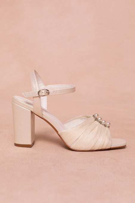 The Piper Pearl Heel in Ivory by Ivy City Co features a cream-colored design with a block pearl heel, ankle strap and buckle, and pearl accents on the front—ideal ivory heels for special occasions. Set against a soft beige background.