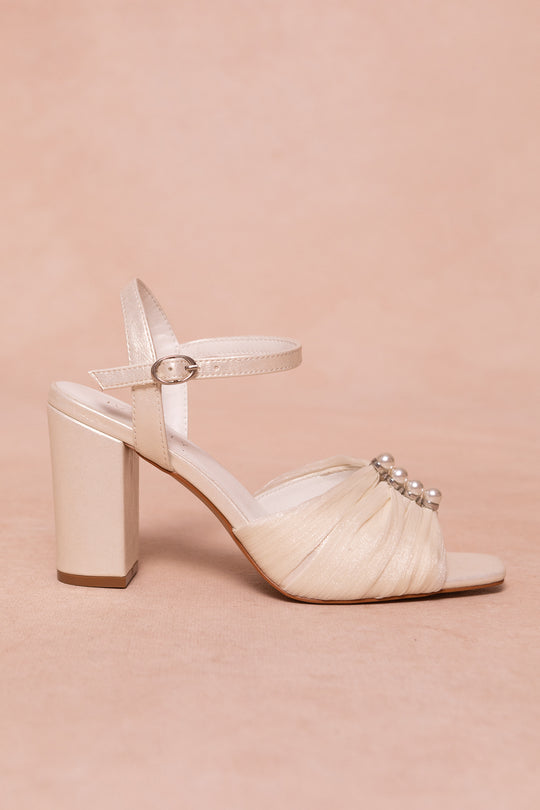 The Piper Pearl Heel in Ivory by Ivy City Co features a cream-colored design with a block pearl heel, ankle strap and buckle, and pearl accents on the front—ideal ivory heels for special occasions. Set against a soft beige background.