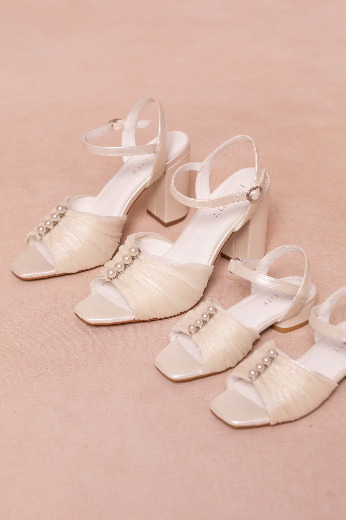 Two pairs of Ivy City Co’s Mini Piper Pearl Heel in Ivory sandals, featuring ankle straps, tulle accents, and pearl embellishments, are displayed on a soft beige surface—perfect vegan leather girls dress shoes.