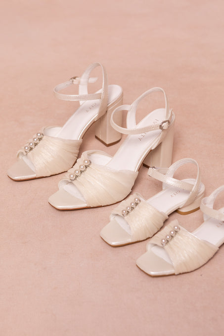 Two pairs of Ivy City Co’s Mini Piper Pearl Heel in Ivory sandals, featuring ankle straps, tulle accents, and pearl embellishments, are displayed on a soft beige surface—perfect vegan leather girls dress shoes.