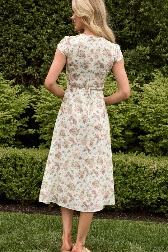 A woman stands barefoot on grass, facing away toward green bushes, wearing the Ivy City Co Fiona Dress in Countryside Peony Blossom—a light-colored, floral midi dress with short sleeves and a tie at the back.