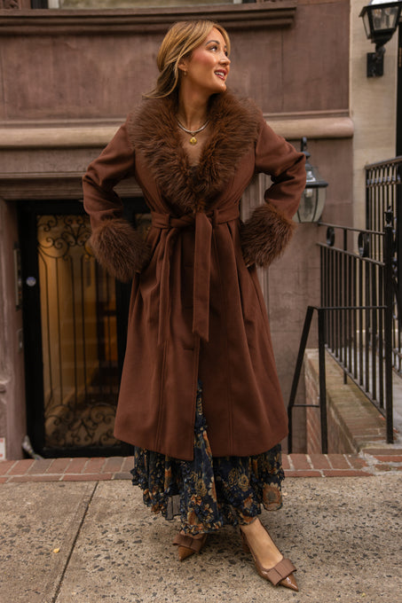 A woman stands outdoors on a city sidewalk by the stairs, smiling in the Ivy City Co Pookie Coat in Warm Cocoa. The long coat features fur trim on the collar and cuffs, which she pairs with a floral dress and pointed brown shoes.