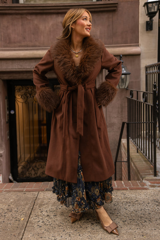A woman stands outdoors on a city sidewalk by the stairs, smiling in the Ivy City Co Pookie Coat in Warm Cocoa. The long coat features fur trim on the collar and cuffs, which she pairs with a floral dress and pointed brown shoes.