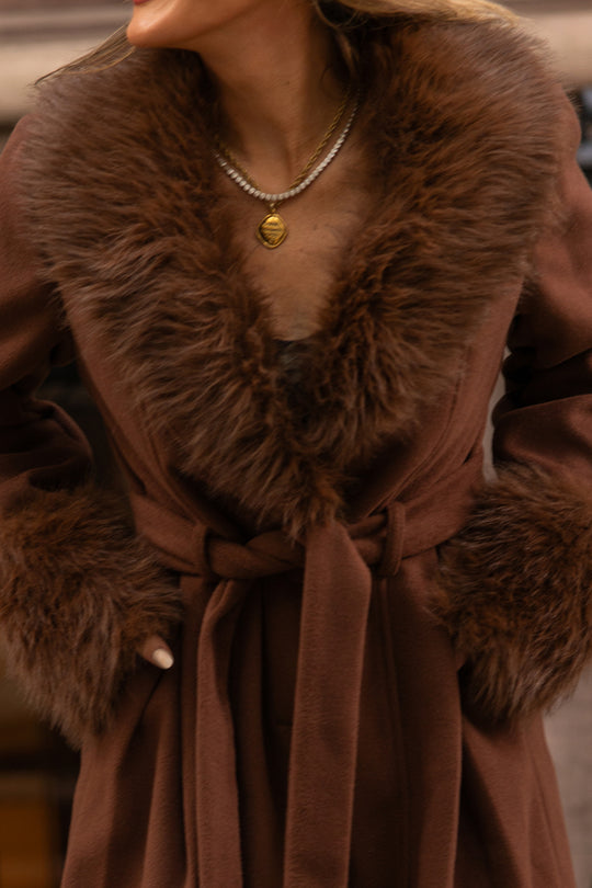 Wearing the Ivy City Co Pookie Coat in Warm Cocoa, which features a thick fur collar and cuffs, hands tucked into its pockets and accessorized with a gold pendant necklace and a strand of pearls. The person’s face is partially out of frame.