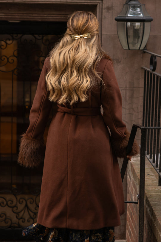 A woman with long, wavy blonde hair, styled with a gold hair clip, wears the Ivy City Co Pookie Coat in Warm Cocoa—featuring plush fur cuffs—while standing outdoors near a building entrance, facing away from the camera.