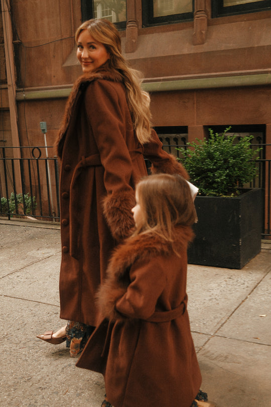 A woman and a young girl, both smiling, walk hand in hand on a city sidewalk wearing matching Ivy City Co Pookie Coats in Warm Cocoa with fur trim.