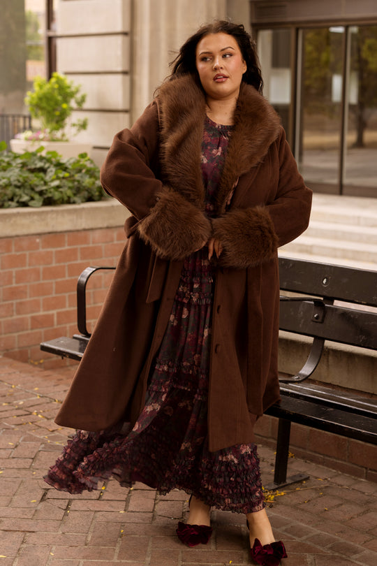 A woman wearing the Ivy City Co Pookie Coat in Warm Cocoa with fur trim and a floral dress walks by a black bench on a brick path, near a brick wall and green plants, with buildings in the background.