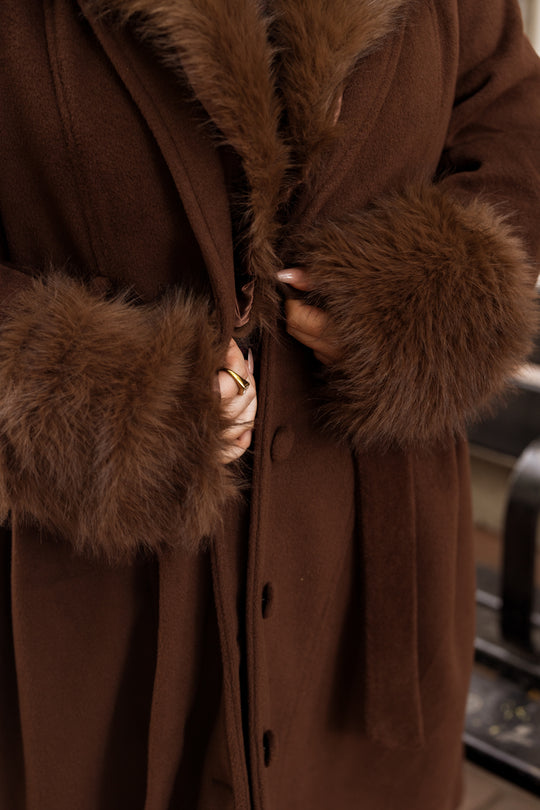 A person is buttoning up the Ivy City Co Pookie Coat in Warm Cocoa, featuring fluffy fur cuffs and collar. Only their hands and the cozy brown coat are visible in the image.