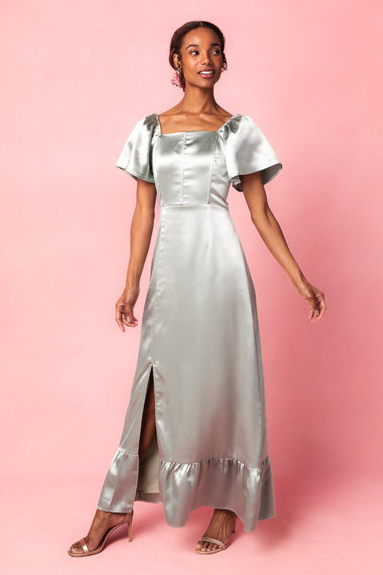 A woman wearing the Maddie Dress in Sage Satin by Ivy City Co—a shiny sage green maxi event dress with short flutter sleeves, a square neckline, front buttons, and a front slit—standing on a pink background.