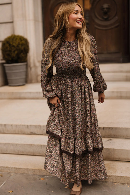 A woman with long hair, wearing the Presley Dress in Ditsy Autumn Fleur by Ivy City Co, stands on stone steps outdoors, smiling and looking to the side.