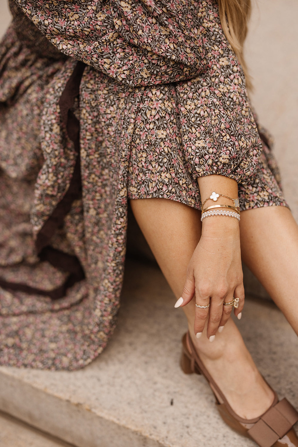 A woman wearing the Presley Dress in Ditsy Autumn Fleur by Ivy City Co sits on stone steps with her legs crossed. She accessorizes with gold bracelets, rings, brown shoes, and neutral-toned nail polish.