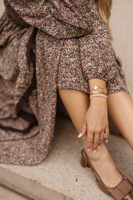 A woman wearing the Presley Dress in Ditsy Autumn Fleur by Ivy City Co sits on stone steps with her legs crossed. She accessorizes with gold bracelets, rings, brown shoes, and neutral-toned nail polish.