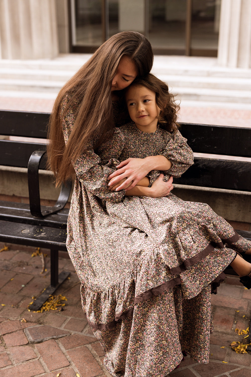 A woman and a young girl, both wearing Ivy City Co’s Presley Dress in Ditsy Autumn Fleur, sit together on a bench. The woman hugs the girl closely as they sit calmly among scattered yellow leaves.