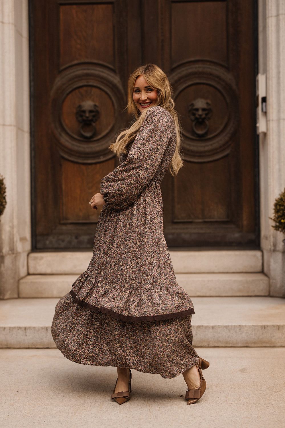 A woman wearing the Presley Dress in Ditsy Autumn Fleur by Ivy City Co smiles as she twirls on stone steps before grand, ornate wooden doors. She pairs her long, patterned dress with heeled shoes and lets her wavy blonde hair cascade down, perfectly embodying the autumnal elegance of this Ivy City Co favorite.