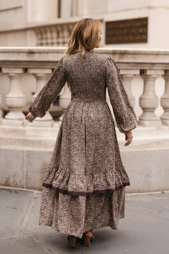 A woman with long, wavy hair stands outdoors with her back to the camera near a stone railing, wearing the Ivy City Co Presley Dress in Ditsy Autumn Fleur—a floral-patterned, long-sleeved dress—paired with heeled shoes.