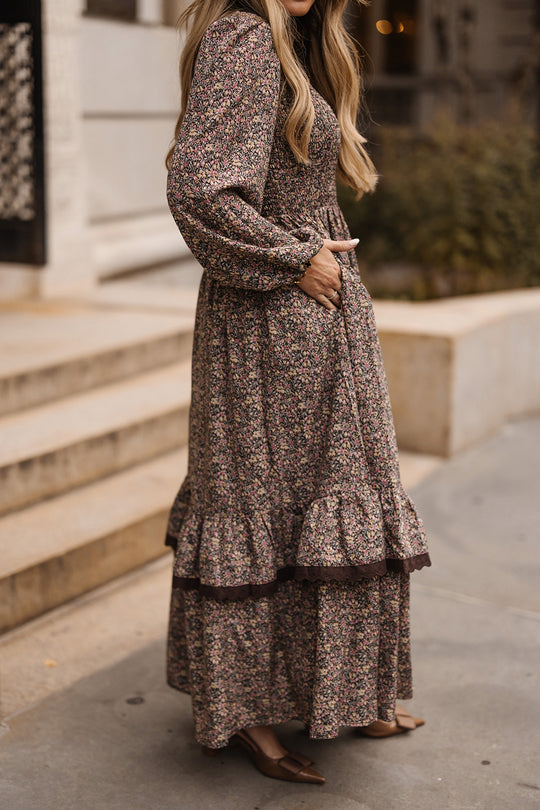 A woman stands on a sidewalk, hands in pockets, wearing Ivy City Co’s Presley Dress in Ditsy Autumn Fleur—its long sleeves, ruffled hem, and floral maxi silhouette offering a perfect blend of comfort and fall style. She has long, wavy blonde hair. The photo is cropped just above her head.
