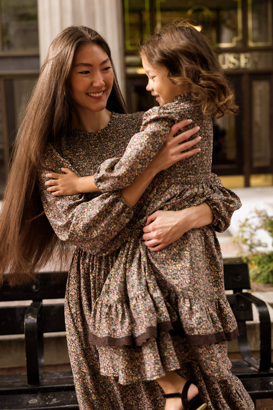 A woman with long hair smiles while holding a young girl. Both wear matching Ivy City Co Presley Dresses in Ditsy Autumn Fleur and stand outdoors in front of a building.