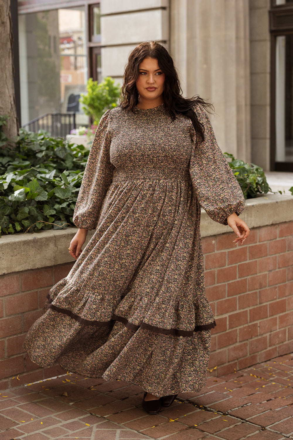 A woman wearing the Ivy City Co Presley Dress in Ditsy Autumn Fleur, with its long sleeves and floral pattern, stands outside by a brick wall and green plants, looking confidently toward the camera.