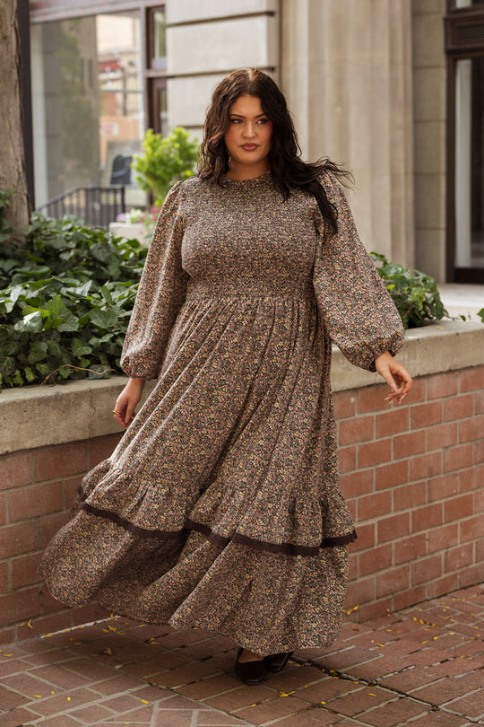 A woman wearing the Ivy City Co Presley Dress in Ditsy Autumn Fleur, with its long sleeves and floral pattern, stands outside by a brick wall and green plants, looking confidently toward the camera.