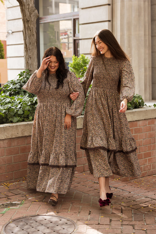 Two women wearing the Ivy City Co Presley Dress in Ditsy Autumn Fleur walk together outside, smiling and holding hands next to a brick planter filled with greenery and trees, in front of a building with large windows and columns.