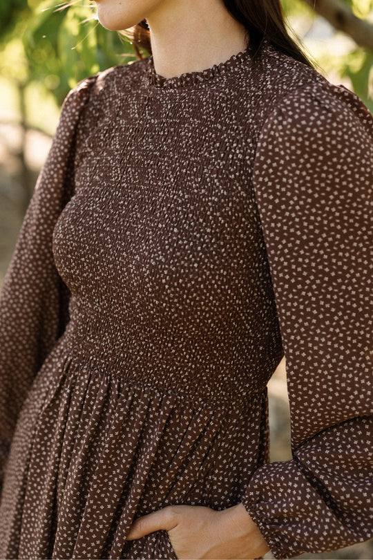 A woman stands outdoors among trees, her upper body visible and one hand tucked in her pocket, wearing the Ivy City Co Presley Dress in Mahogany Ditsy—a brown long-sleeve dress with a small white print and smocked bodice.