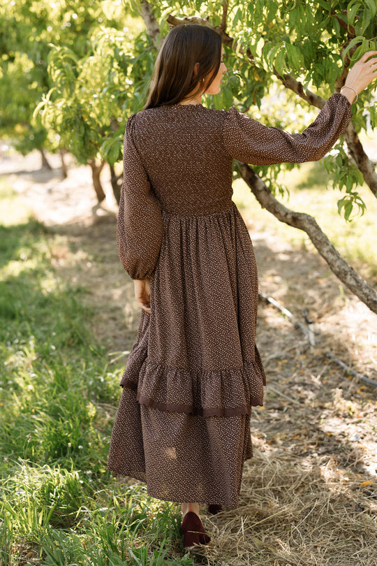 A woman in the Ivy City Co Presley Dress in Mahogany Ditsy, featuring a smocked bodice and elegant balloon sleeves, stands outdoors with her back to the camera, gently touching a leafy tree branch in a sunlit orchard.