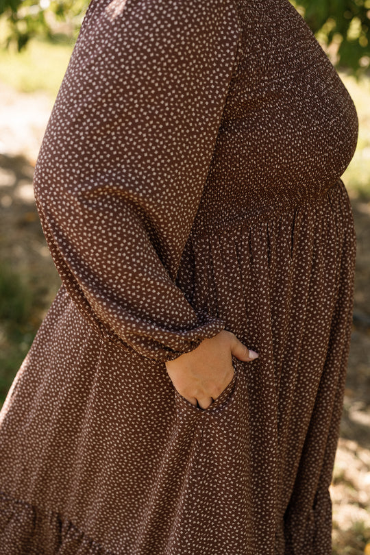 A person wearing the Ivy City Co Presley Dress in Mahogany Ditsy, featuring long sleeves, white polka dots, and stylish balloon sleeves, stands outdoors with one hand in their pocket. The background is blurred with sunlight and greenery.