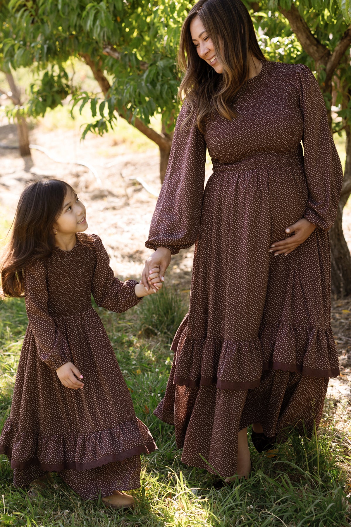A woman and a young girl, both dressed in matching Ivy City Co Presley Dress in Mahogany Ditsy with smocked bodices and elegant balloon sleeves, hold hands and smile at each other while standing outdoors among green trees and grass.