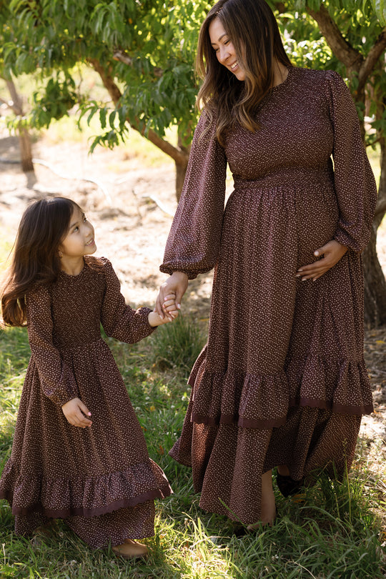 A woman and a young girl, both dressed in matching Ivy City Co Presley Dress in Mahogany Ditsy with smocked bodices and elegant balloon sleeves, hold hands and smile at each other while standing outdoors among green trees and grass.