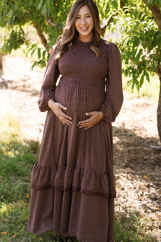 A pregnant woman in the Ivy City Co Presley Dress in Mahogany Ditsy, featuring balloon sleeves and a smocked bodice, stands outdoors smiling and gently cradling her belly with both hands. Sunlight filters through green trees in the background.