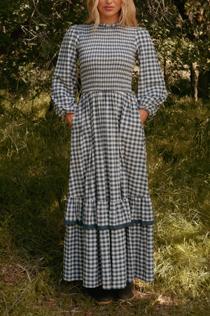 A woman stands on grass wearing the Ivy City Co Presley Dress in Indigo Gingham, featuring long sleeves, a smocked bodice, ruffled hem, and pockets. Her face is partially out of frame.