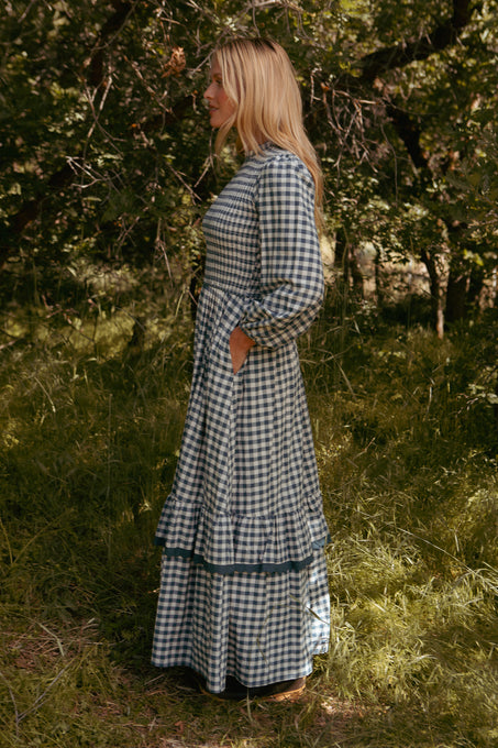 A woman stands in profile in a grassy, wooded area, wearing the Ivy City Co Presley Dress in Indigo Gingham featuring a smocked bodice, ruffled hem, and pockets as sunlight filters through the trees.
