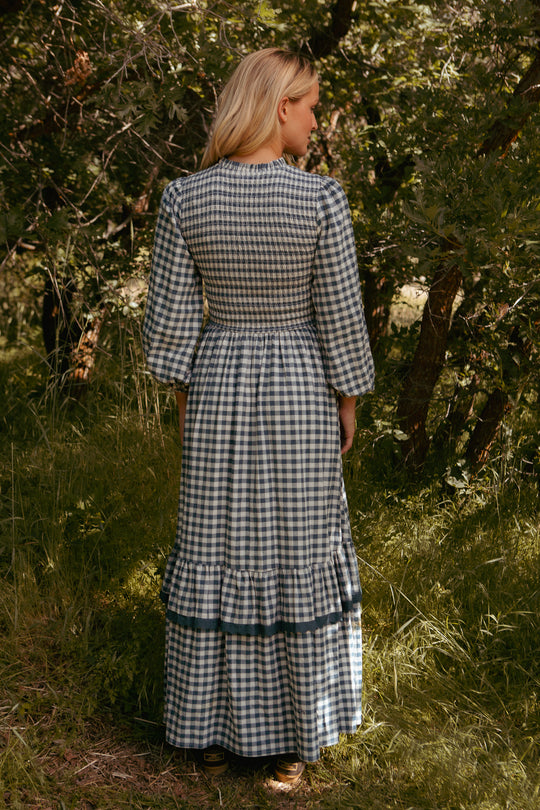 A woman stands outdoors with her back to the camera, wearing the Ivy City Co Presley Dress in Indigo Gingham, which features ruffle details and a smocked bodice. She is surrounded by lush green trees and grass.