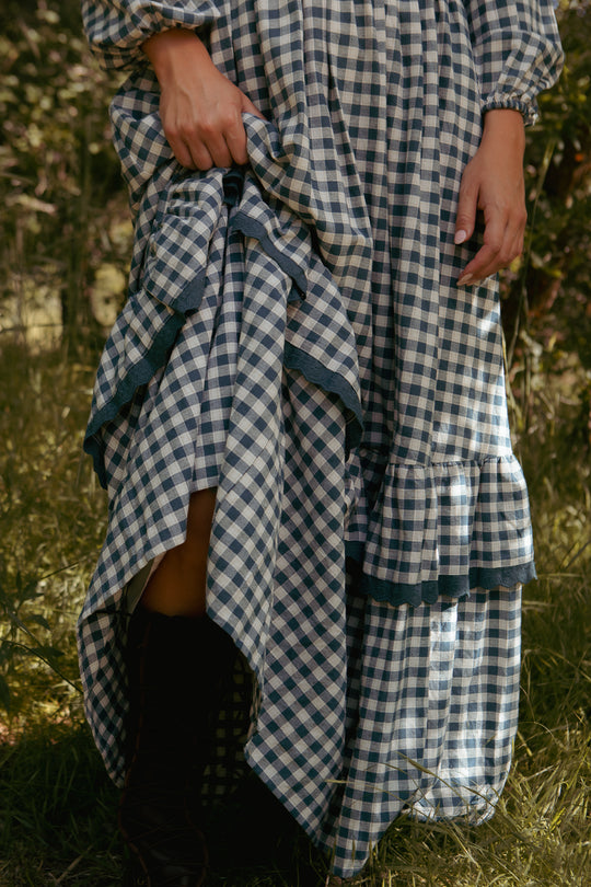 A person in the Ivy City Co Presley Dress in Indigo Gingham stands outdoors, lifting the maxi dress to reveal dark boots beneath. The sunlit, grassy setting adds a welcoming touch.