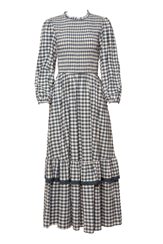 The Presley Dress in Indigo Gingham by Ivy City Co features a smocked bodice, long sleeves, ruffled neckline, and a tiered ankle-length skirt with gathered details.
