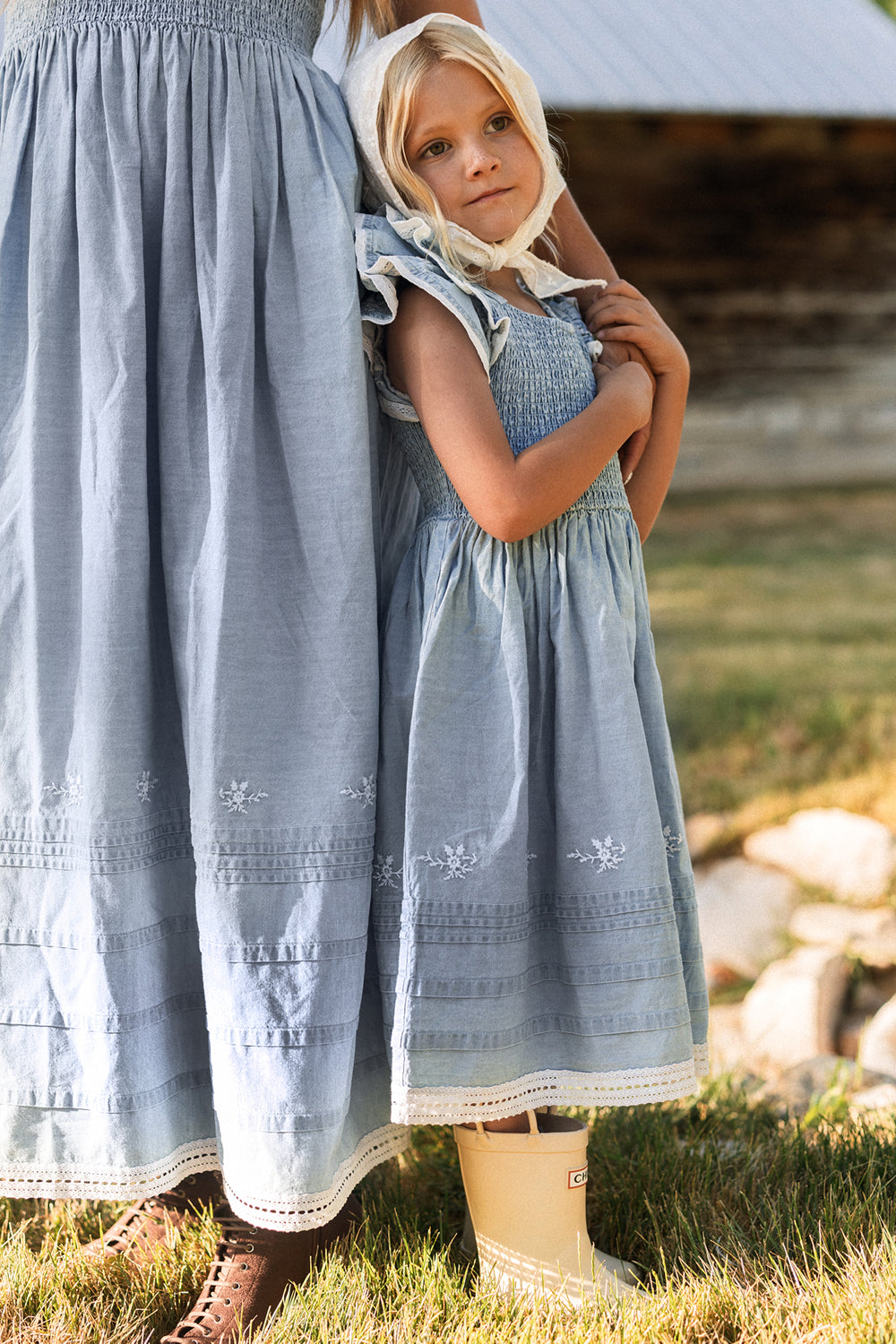 A young girl in a light blue dress with a smocked bodice and cream rain boots stands outside, holding onto an adult wearing the Ivy City Co Hattie Ankle Length Dress in Embroidered Chambray, which is maternity friendly. They are on a grassy lawn with a rustic wooden building behind them.