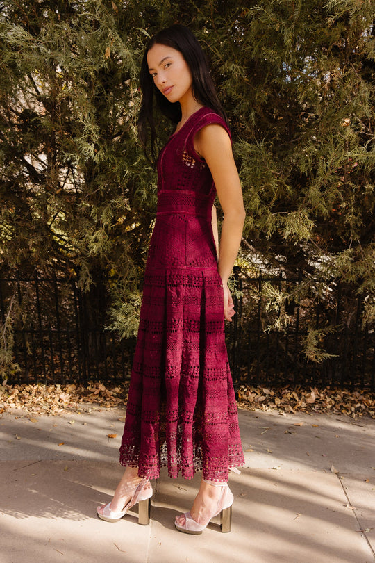 A woman wearing the Amory Dress in Burgundy by Ivy City Co—a sleeveless lace midi dress with a square neckline—pairs it with pink high heels as she stands on a sunlit sidewalk near leafy bushes and a black fence, gazing softly at the camera.