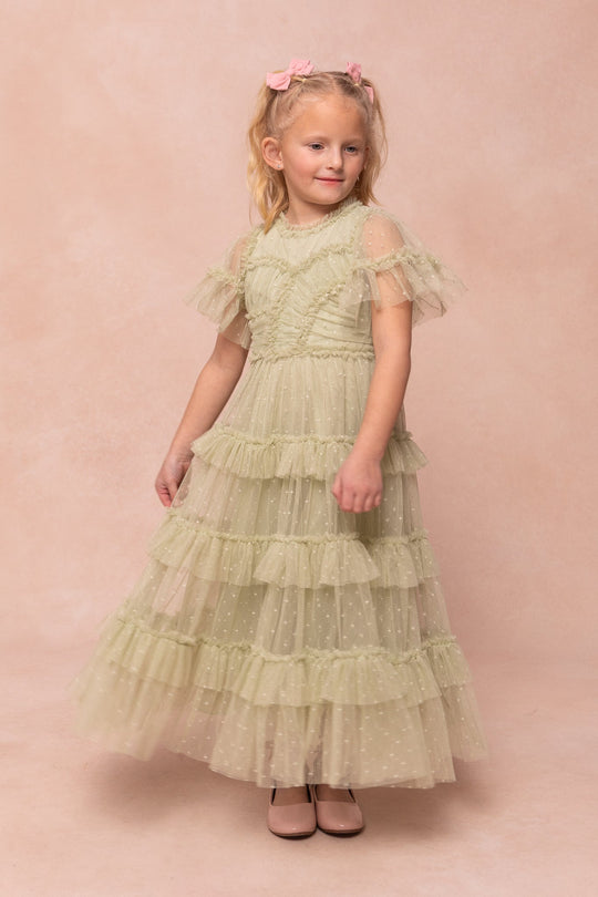 A young girl stands against a soft pink background, smiling slightly as she models the Ivy City Co Mini Whimsical Maxi Dress in Sage. The pale green, ruffled tulle dress features sheer sleeves and falls to maxi length. Her blonde hair is styled with two pink bows, and she wears light pink shoes that perfectly complement her twirl-worthy outfit.
