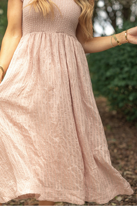 A woman stands outdoors amid green foliage, wearing the Ivy City Co Hattie Dress in Pink Stripes—a textured midi dress with a smocked bodice. She holds the skirt with one hand, her long, wavy blonde hair cascading over her shoulder and gold bracelets adorning her wrist.