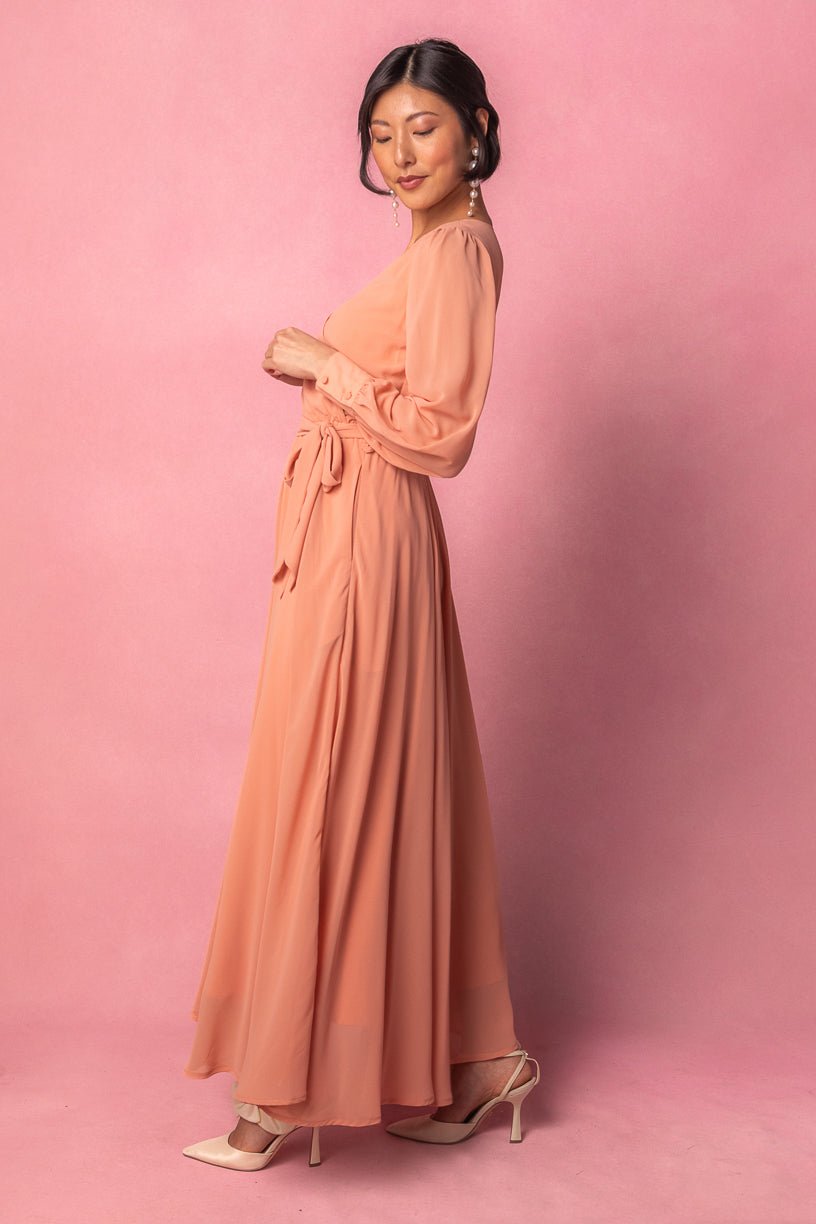 A woman stands sideways against a pink background, wearing the Ivy City Co Andie Dress in Apricot Crush—a long maxi dress with long sleeves and a waist tie—paired with beige heels. Her hair is styled up, and she accessorizes with long pearl earrings.