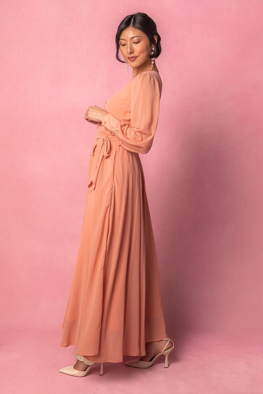 A woman stands sideways against a pink background, wearing the Ivy City Co Andie Dress in Apricot Crush—a long maxi dress with long sleeves and a waist tie—paired with beige heels. Her hair is styled up, and she accessorizes with long pearl earrings.
