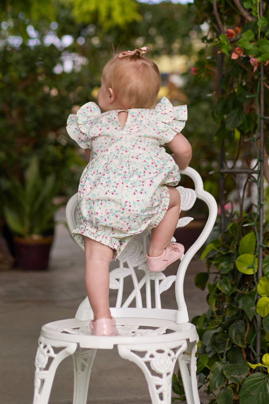 A baby wearing the Ivy City Co Baby Hattie Romper in Pansy Trellis climbs onto a white ornate chair, surrounded by greenery, with her back facing the camera.