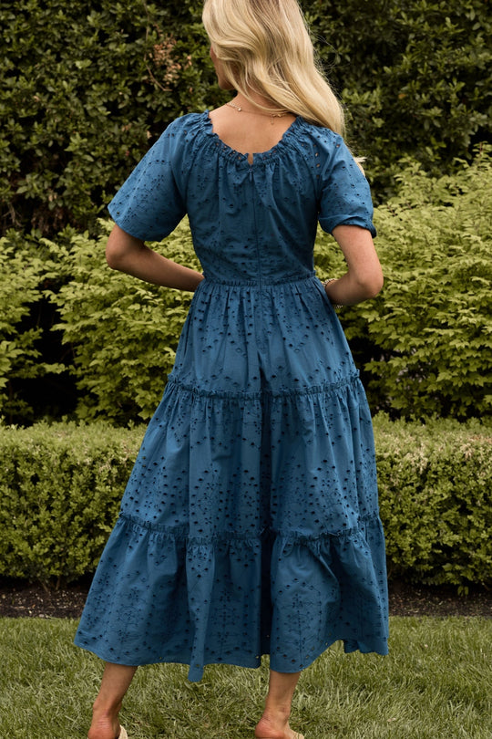 A woman with long blonde hair stands outdoors, facing away, wearing the Etta Dress in Cobalt by Ivy City Co—a summer midi dress with an eyelet pattern and short sleeves in a striking cobalt blue. Lush green bushes and grass are in the background.