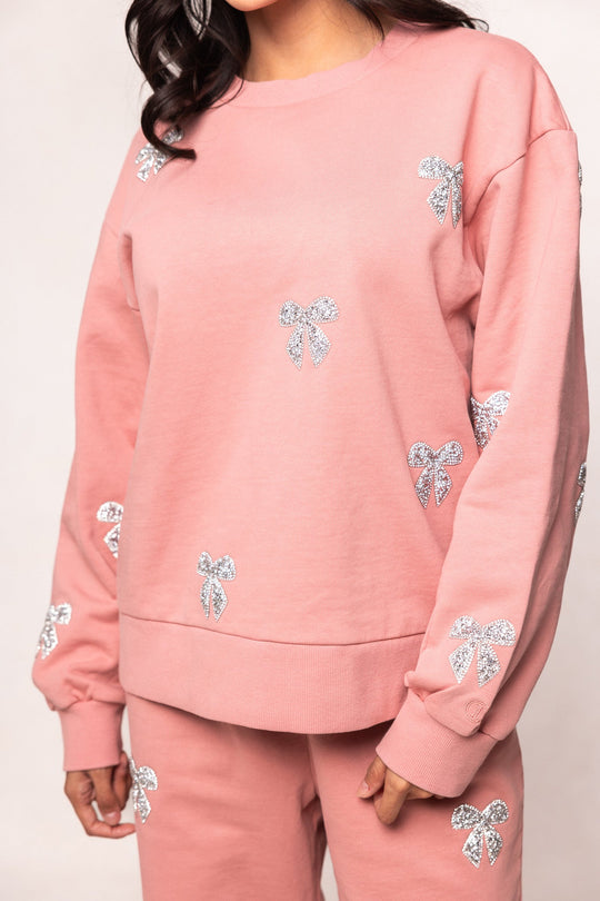 A person is wearing the Ivy City Co Micah Crew Neck Sweatshirt in Pink & Bows along with matching lounge pants, both featuring silver glitter bows from the Ivy City Co Lounge Collection. The person’s face is not visible in the image.