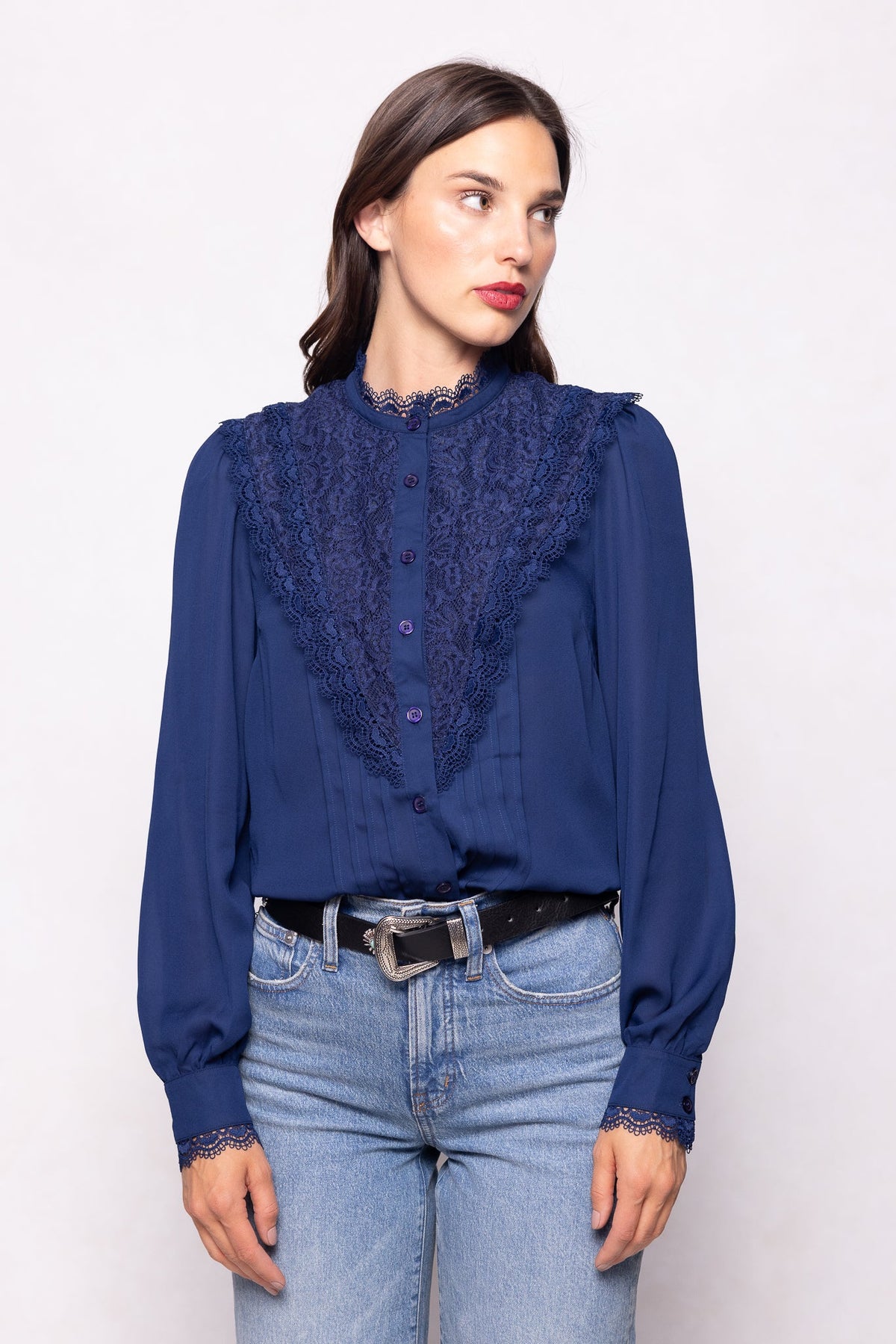 A woman, standing against a plain light background, wears the Ivy City Co Blythe Blouse in Navy—featuring long sleeves, lace details, and buttons for a stylish and nursing-friendly touch—tucked into light blue jeans with a black belt.