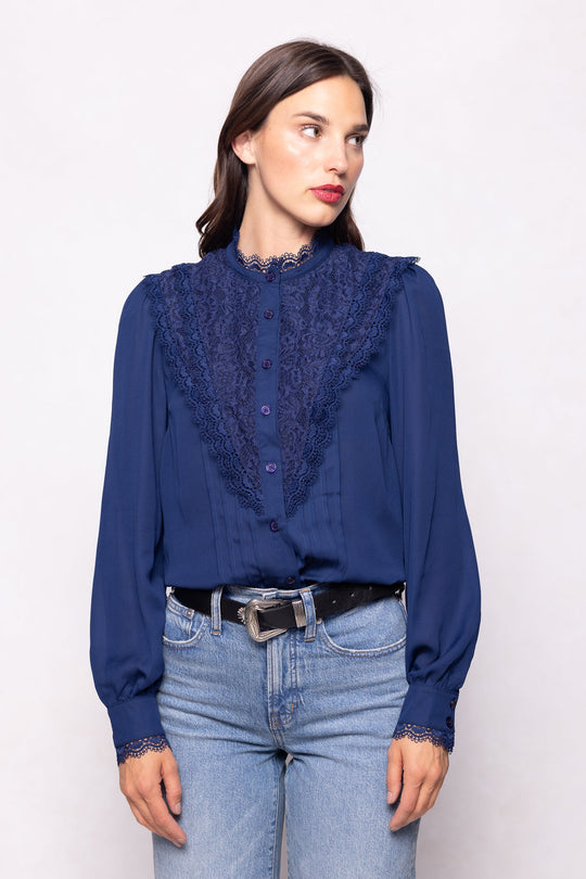 A woman, standing against a plain light background, wears the Ivy City Co Blythe Blouse in Navy—featuring long sleeves, lace details, and buttons for a stylish and nursing-friendly touch—tucked into light blue jeans with a black belt.