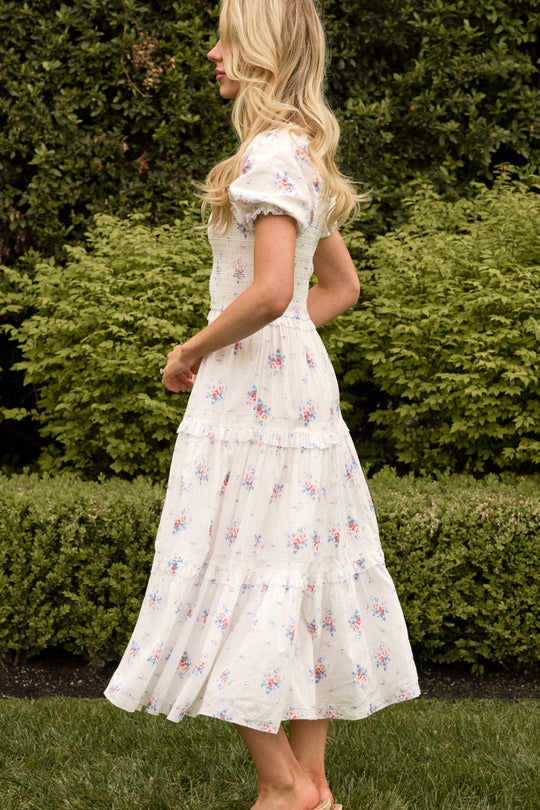 A woman with long blonde hair stands barefoot in profile on grass, wearing the Ivy City Co Emily Dress in Heritage Soft Ditsy Floral—a light, short-sleeved midi dress with a tiered design. This dress is both nursing friendly and perfect for maternity wear. Green bushes and foliage serve as the backdrop.