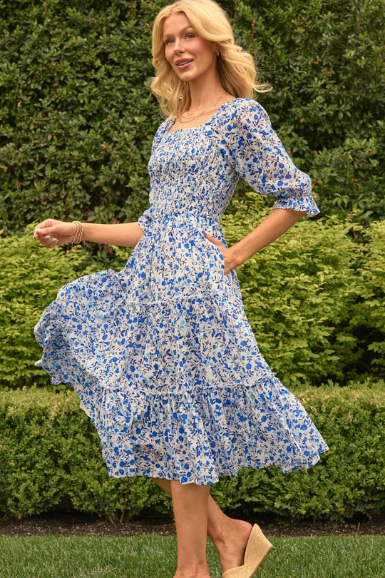 A woman stands outdoors on grass, smiling as she holds the skirt of her Ivy City Co Madeline Dress in Ditsy Cobalt Floral. She accessorizes with wedge sandals and gold bracelets, framed by a backdrop of green shrubs.
