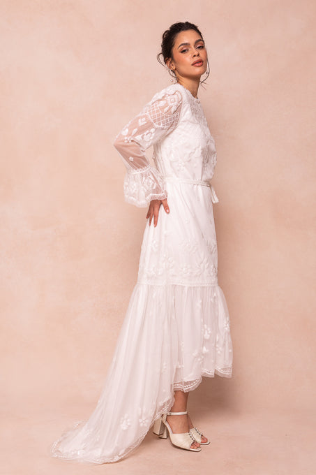 A woman stands sideways in the Ivy City Co Queenie Dress in White, a long tulle gown with sheer embroidered sleeves and a hand-embellished sequin train, paired with white open-toed heels against a soft beige background.